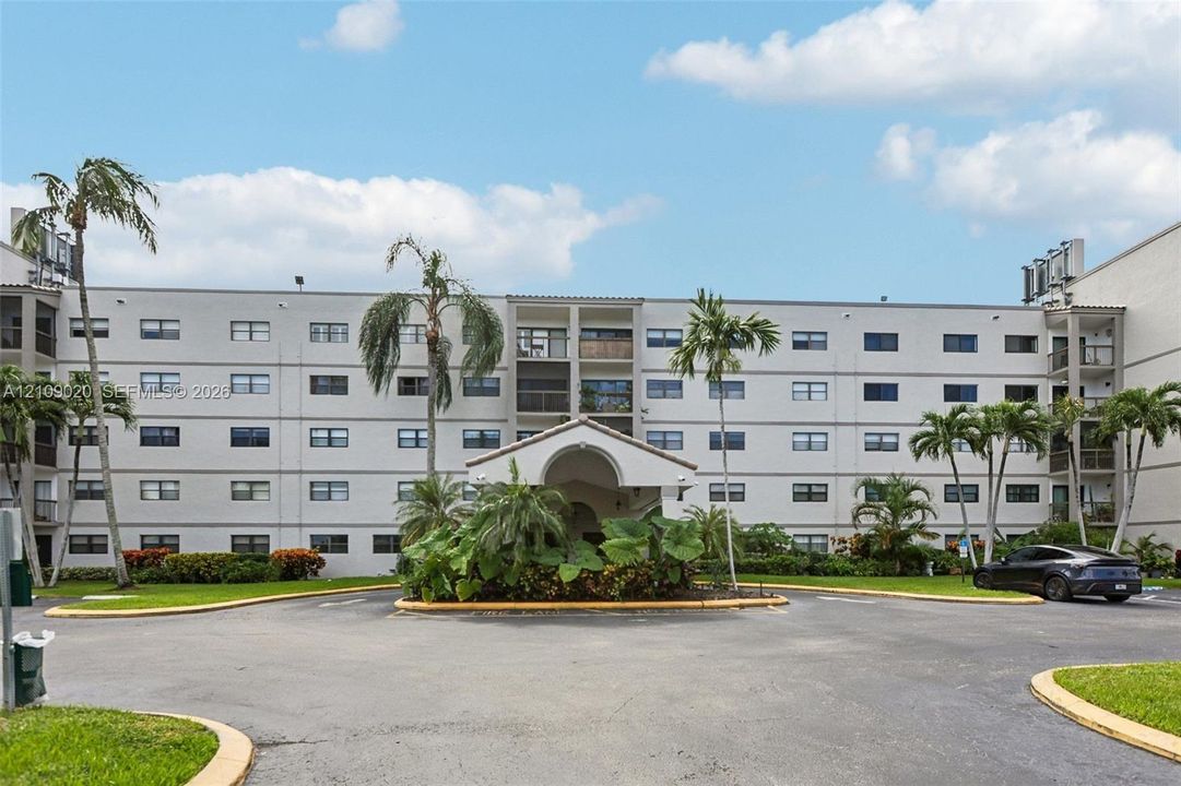 Multi-story stucco exterior featuring a covered entryway, private balconies, and landscaped grounds with mature palm trees