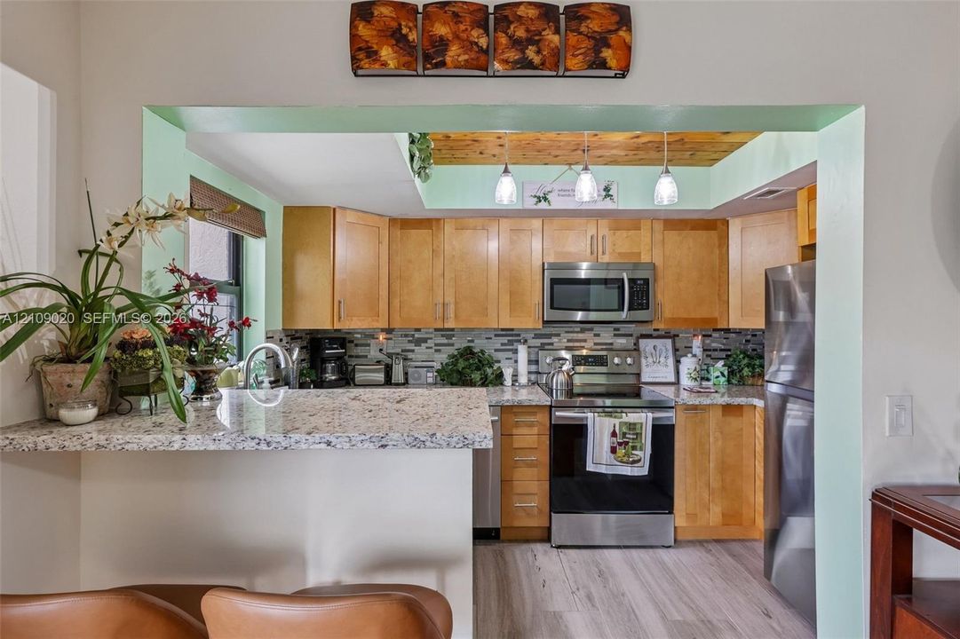 Kitchen featuring wood cabinetry, stone-finish countertops, stainless steel appliances, multi-toned tile backsplash, and recessed wood-plank ceiling detail