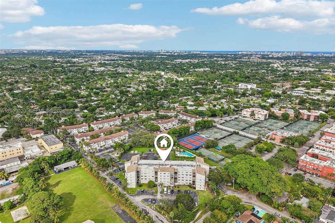Aerial perspective of a multi-story residential building featuring on-site tennis courts, a community pool, surface parking, and extensive mature landscaping