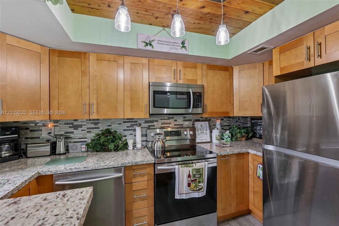Kitchen featuring shaker-style wood cabinetry, stainless steel appliances, speckled stone countertops, a multi-tonal tile backsplash, and a wood-plank ceiling with pendant lighting