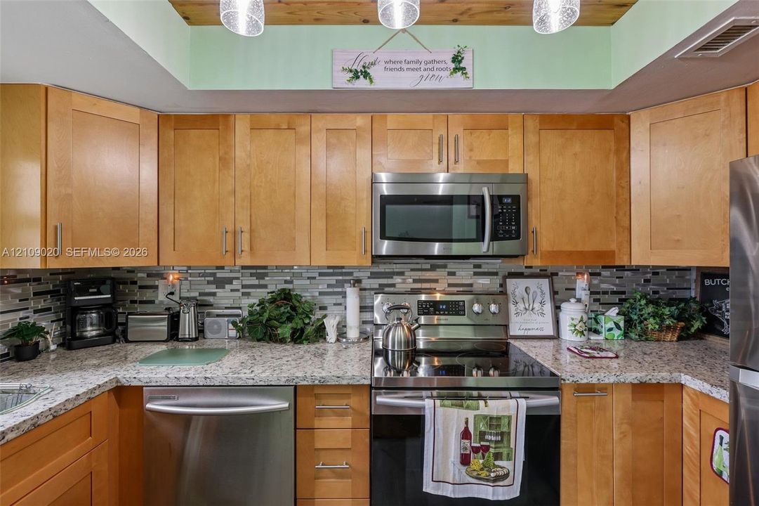 Kitchen featuring natural wood cabinetry, stone-patterned countertops, stainless steel appliances, multi-tonal linear tile backsplash, and wood-paneled tray ceiling