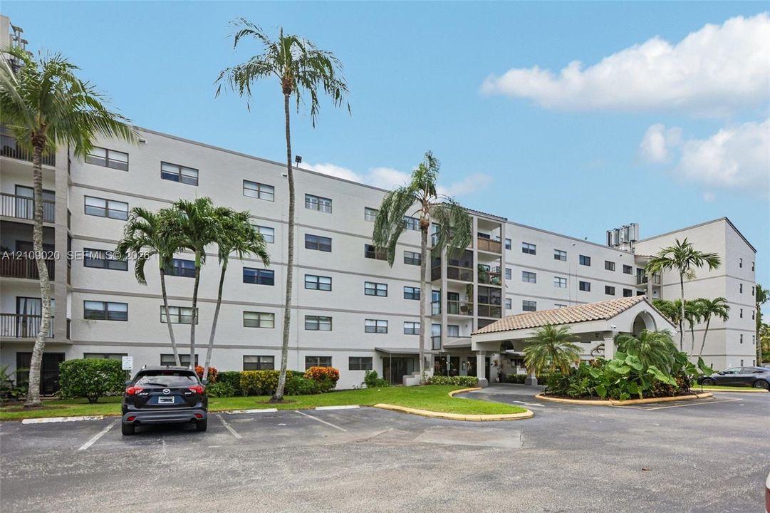 Mid-rise residential building exterior featuring a covered entryway with a tile roof, paved surface parking, and mature palm landscaping