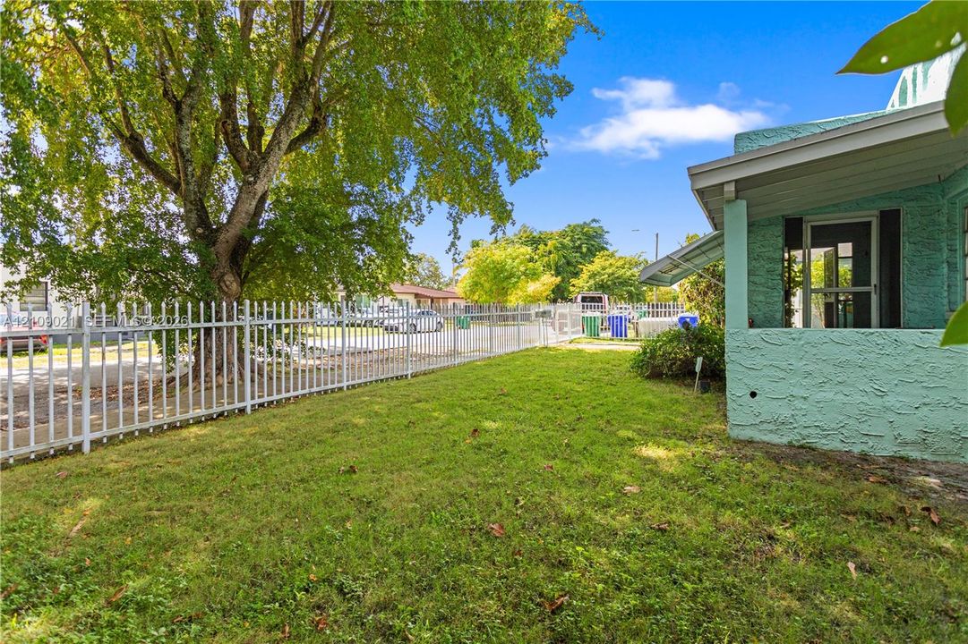 Exterior yard space featuring a metal perimeter fence, mature trees, textured stucco siding, and a covered porch area