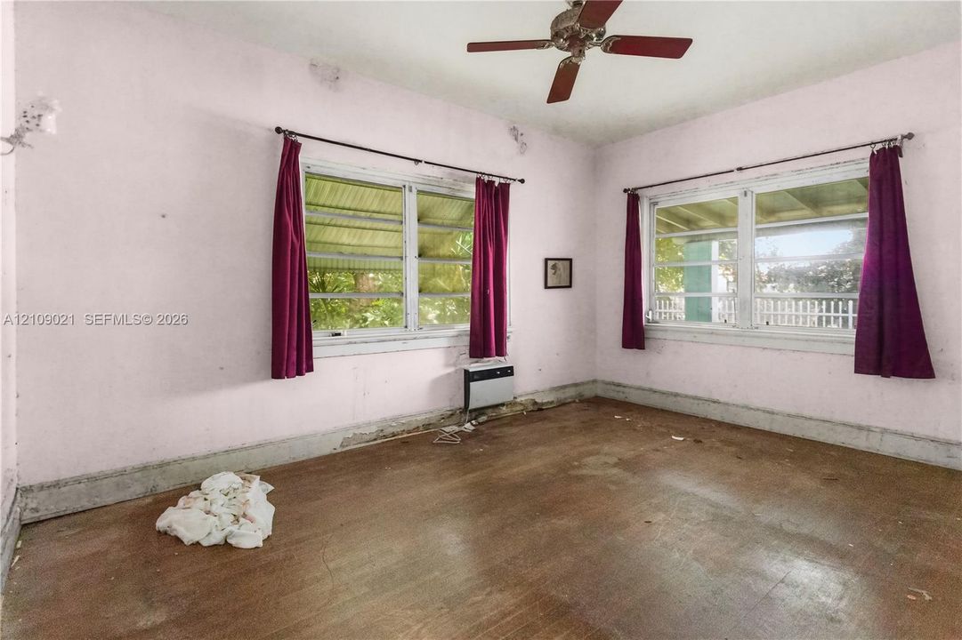 Room featuring wood-finish flooring, two sets of windows with metal-framed screens, a ceiling fan, and a wall-mounted climate control unit