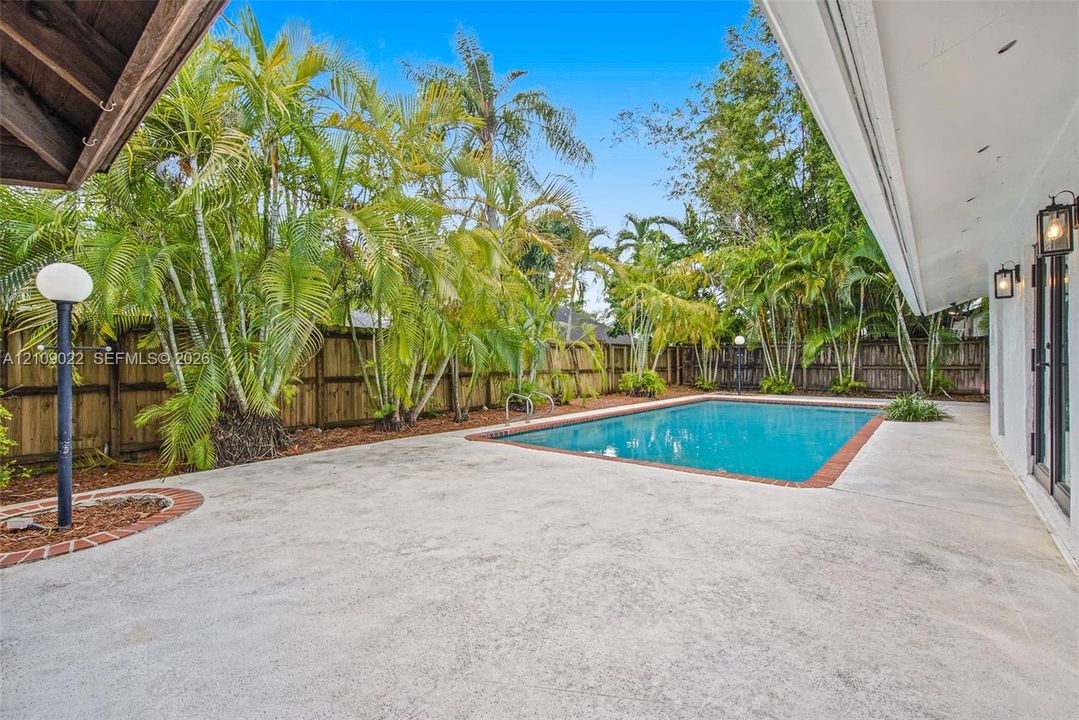 Outdoor space featuring a concrete patio, a swimming pool with a brick-trim border, perimeter wood fencing, and mature palm trees