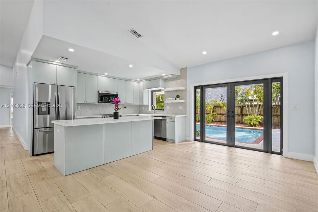 Kitchen featuring light-toned cabinetry, a central island with quartz countertops, stainless steel appliances, geometric tile backsplash, and wood-finish flooring with exterior access to an in-ground pool