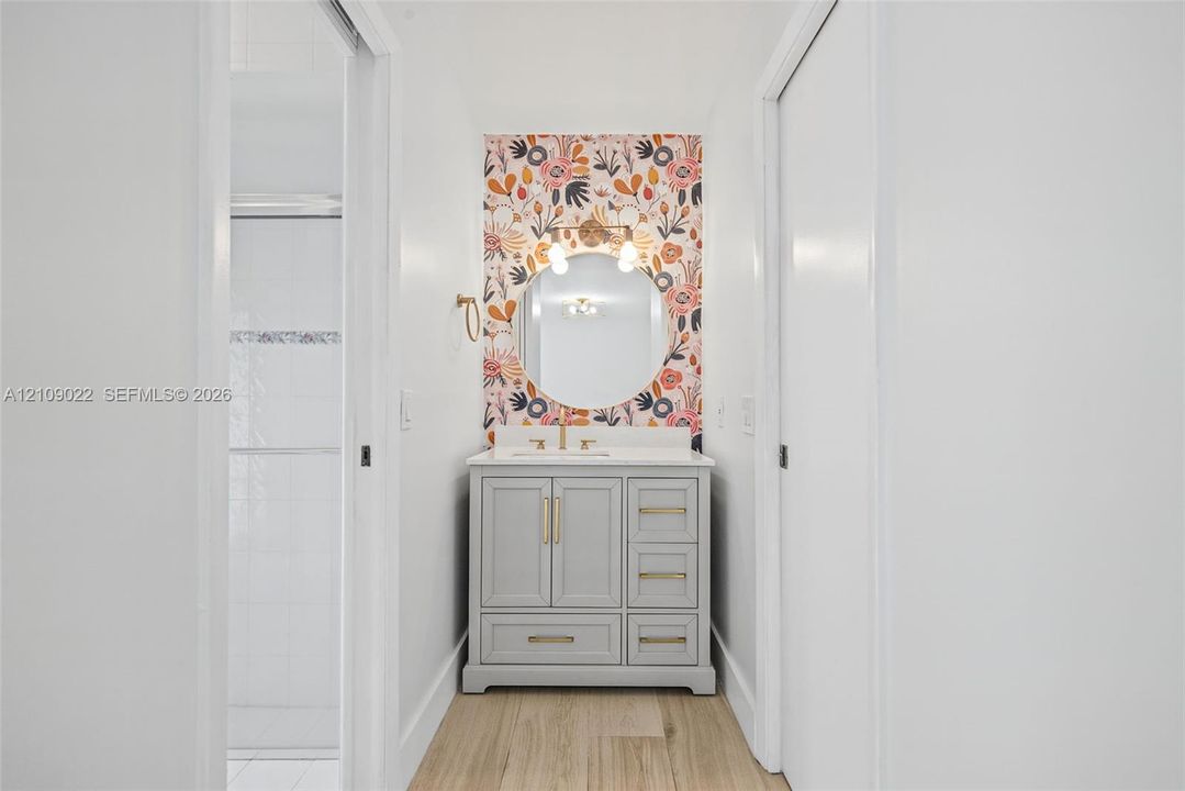 Gray vanity featuring a solid surface countertop, circular wall-mounted mirror, floral patterned wall accent, and metal cabinet hardware