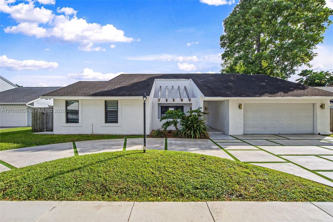 Single-story exterior with white brick facade, dark shingle roof, integrated wooden architectural rafters, multi-car garage, and segmented concrete driveway with inlaid turf