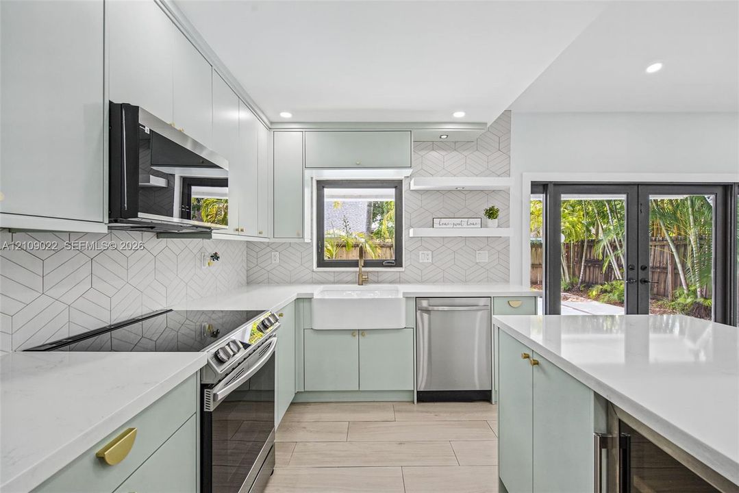 Kitchen featuring soft-toned cabinetry, solid surface countertops, a white farmhouse sink, geometric tile backsplash, and stainless steel appliances