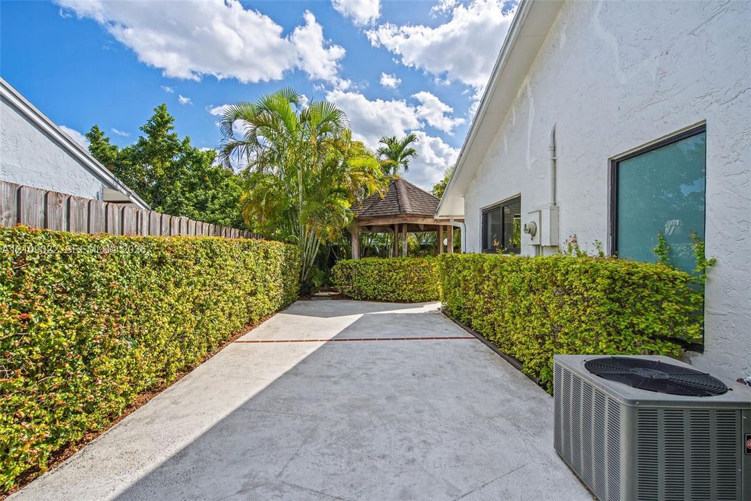 Concrete side yard featuring mature privacy hedging, a wooden gazebo, and textured exterior siding