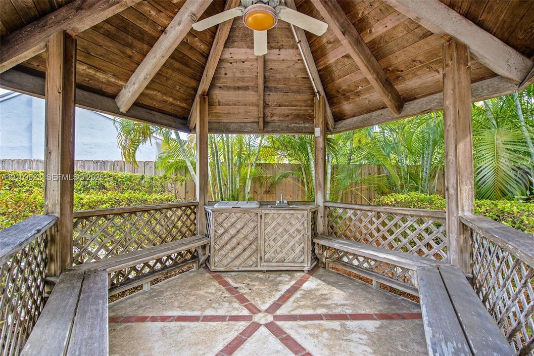 Covered gazebo featuring a wood-plank ceiling, structural timber posts, built-in wooden bench seating, a central ceiling fan, and an integrated outdoor prep station with a sink