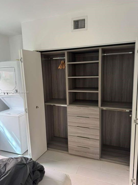Custom wood-finish closet system featuring integrated shelving, four stacked drawers, and multiple hanging rods, accompanied by an in-unit stacked washer and dryer