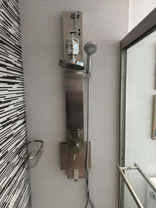Brushed stainless steel shower panel system featuring an integrated rainfall showerhead, adjustable handheld shower wand, and single-handle mixing valve, set against textured wall tile