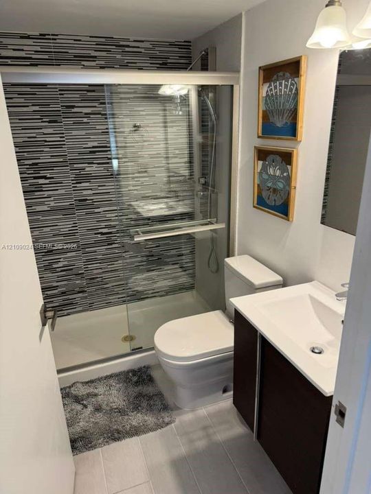Bathroom featuring a walk-in shower with sliding glass doors, decorative horizontal-patterned tile wall surround, dark-finish vanity cabinet, white surface basin, and wall-mounted light fixture