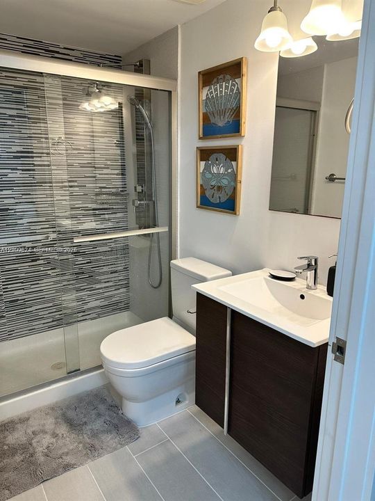 Bathroom featuring a walk-in shower with glass enclosure, multi-toned horizontal tile wall accents, dark wood-finish vanity, integrated sink, and large-format tile flooring