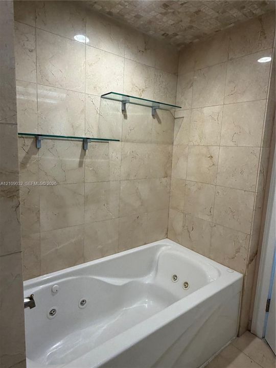 Jetted soaking tub surrounded by floor-to-ceiling stone-tile walls, complemented by dual floating glass shelves and recessed ceiling lighting
