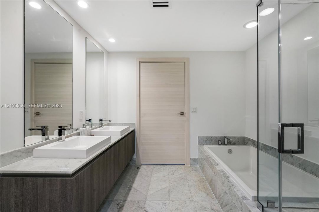 Floating double vanity with dual rectangular vessel sinks, stone-surface countertop, floor-to-ceiling mirrors, recessed lighting, and a glass-enclosed shower with a deep soaking tub