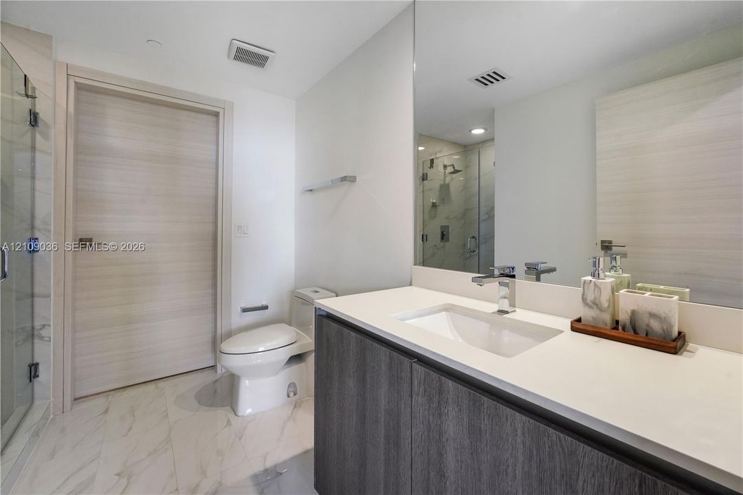 Modern vanity with a light-toned countertop, integrated basin, and dark wood-finish cabinetry, paired with large-format stone-finish floor tile, a full-width mirror, and a glass-enclosed shower