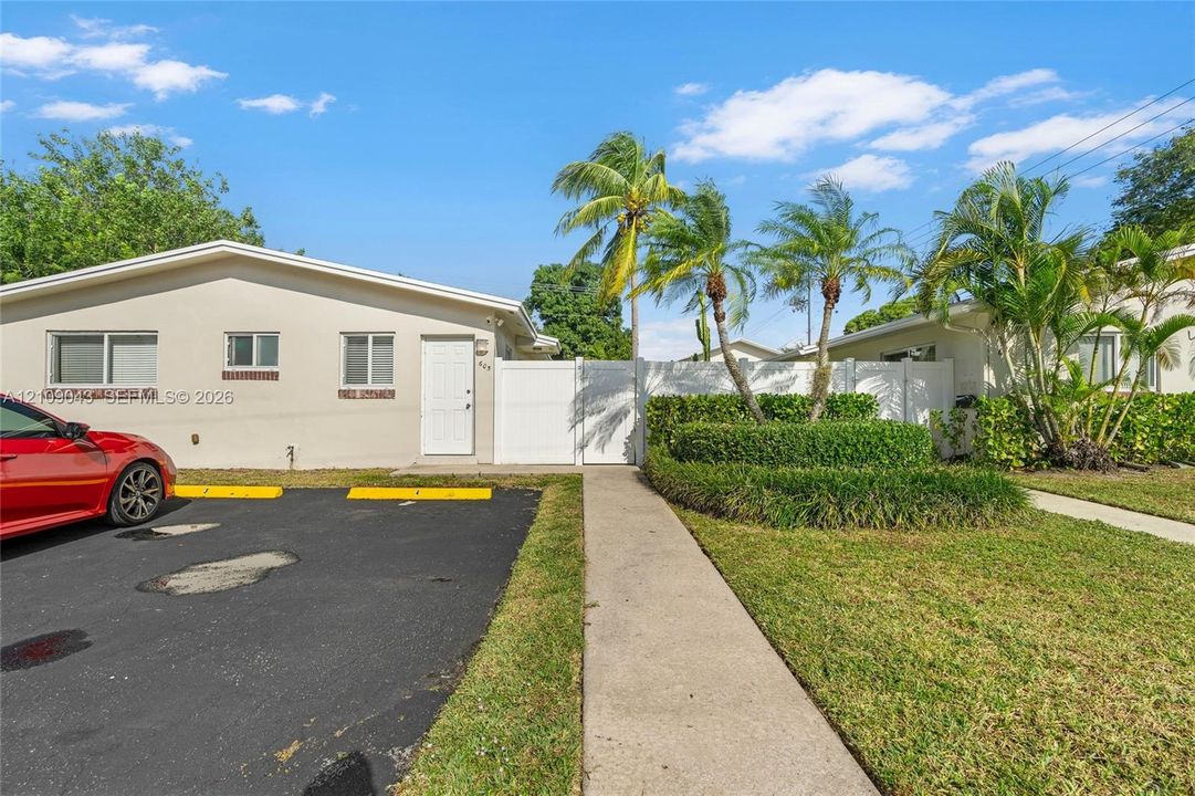 Exterior facade featuring smooth-finish stucco, asphalt parking area, white vinyl privacy fencing, concrete walkway, and landscaped garden beds with palm trees