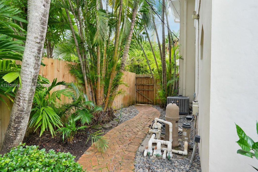 Side yard featuring a brick paver pathway, mature tropical landscaping, and a wood privacy fence with a pedestrian gate