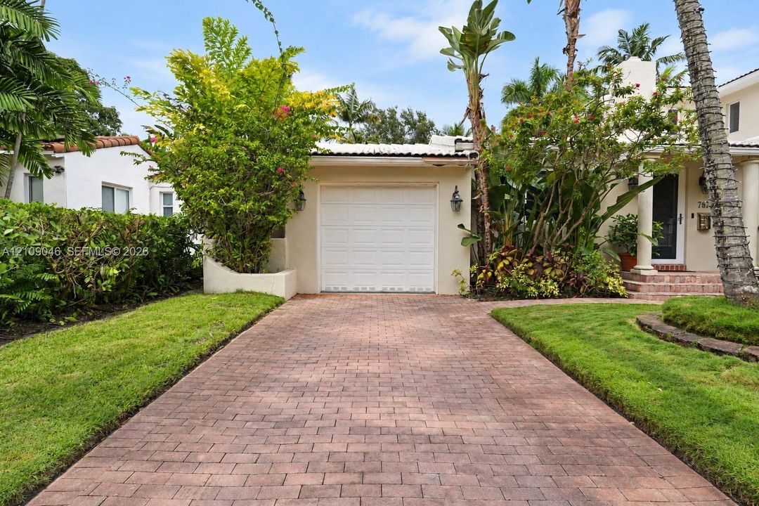 Stucco exterior featuring a single-car garage, brick paver driveway, mature landscaping, and covered entryway with columns