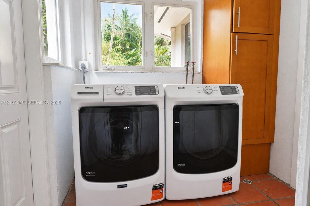 Laundry space featuring floor-to-ceiling wooden cabinetry, terracotta-finish tile flooring, and a window with exterior greenery