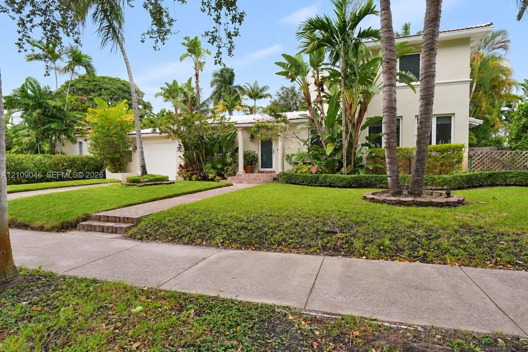 Two-story residence featuring a stucco exterior, a covered entryway with structural columns, brick-paved front steps, and mature tropical landscaping