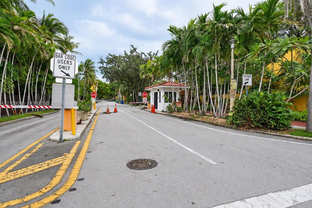 Gated entrance featuring a guardhouse, automated access control, paved thoroughfare, and mature palm landscaping