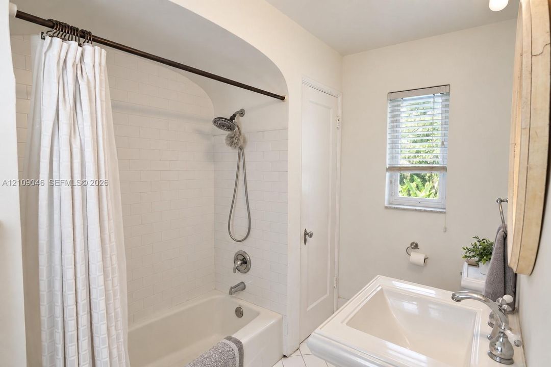 Bathroom featuring a shower-tub combination with white subway tile surround, arched alcove, pedestal sink with metallic fixtures, and a single-hung window with horizontal blinds