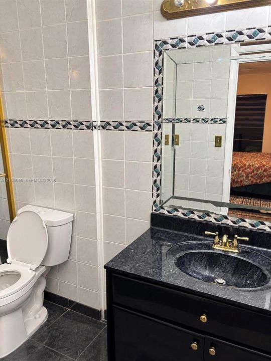 Black vanity cabinet featuring a dark stone-surface countertop, an integrated oval sink, and brass-tone hardware, accented by tiled walls with a geometric-pattern border and a framed wall mirror