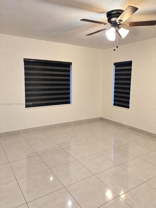 Neutral-toned large format floor tiling, two windows with dual-layer zebra blinds, and a ceiling-mounted fan with integrated multi-bulb lighting