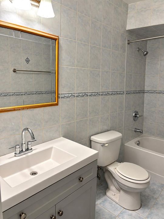 Bathroom featuring a gray vanity with integrated rectangular sink, wall-mounted mirror with a wood-toned frame, tiled walls with decorative accents, and a tub-shower combination