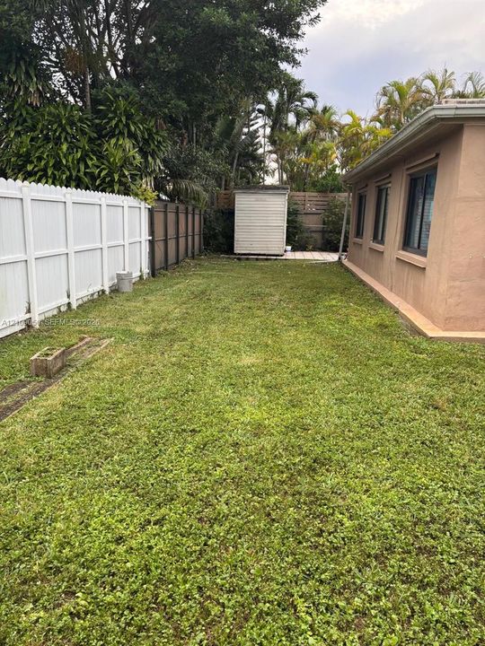 Side yard featuring a grass lawn, white vinyl fencing, mature trees, and a detached storage structure