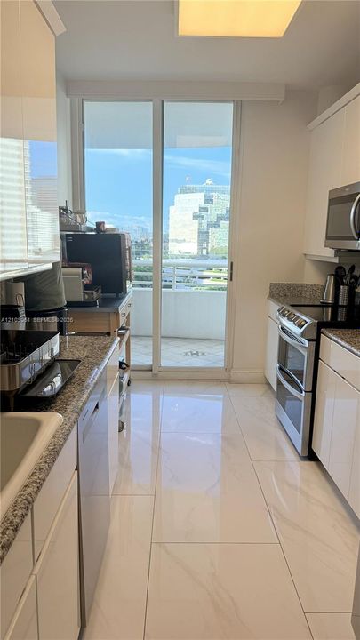 Kitchen featuring high-gloss white cabinetry, stone-finish countertops, stainless steel appliances, large-format tile flooring, and a glass sliding door opening to an exterior balcony