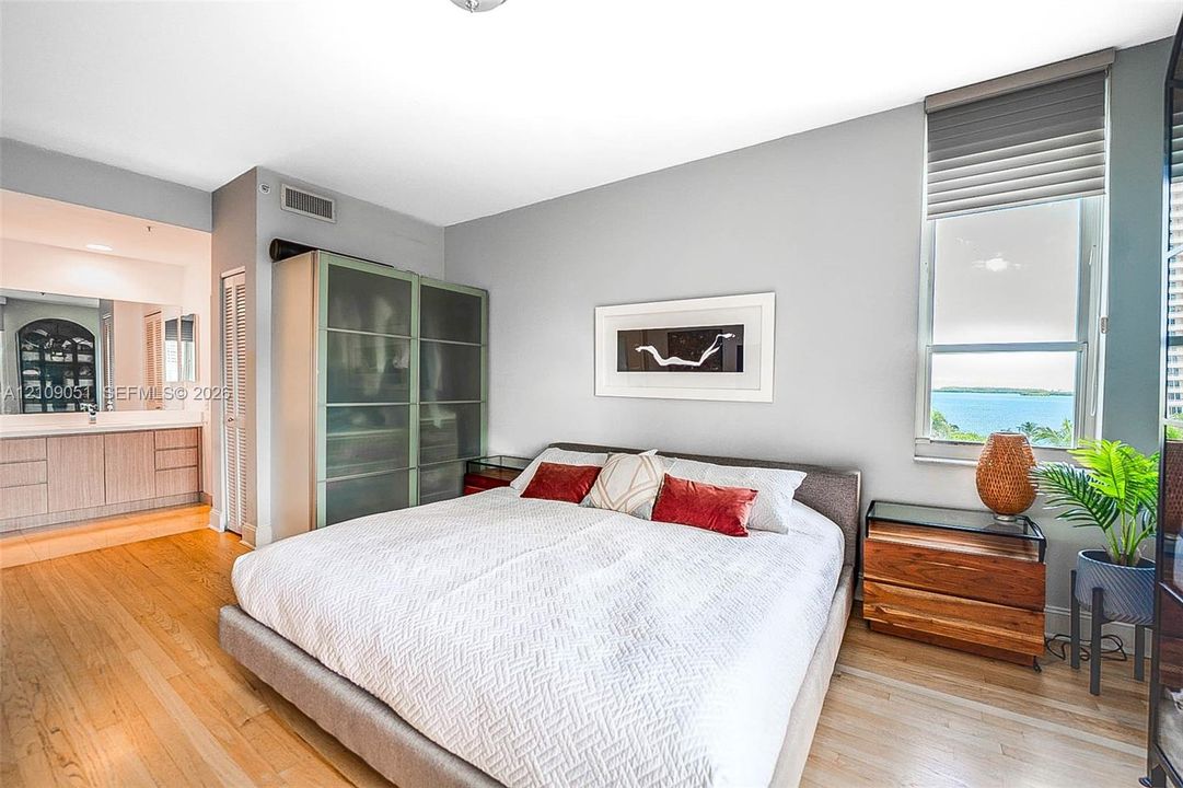 Bedroom featuring wood-finish flooring, floor-to-ceiling frosted glass built-in cabinetry, and a large window with roller shades