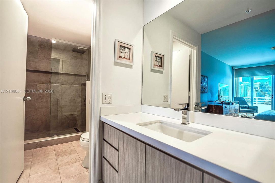 Bathroom featuring a wood-finish vanity with a stone countertop, an integrated rectangular sink, a wall-spanning mirror, and a walk-in shower with a glass enclosure and stone-tile wall surround