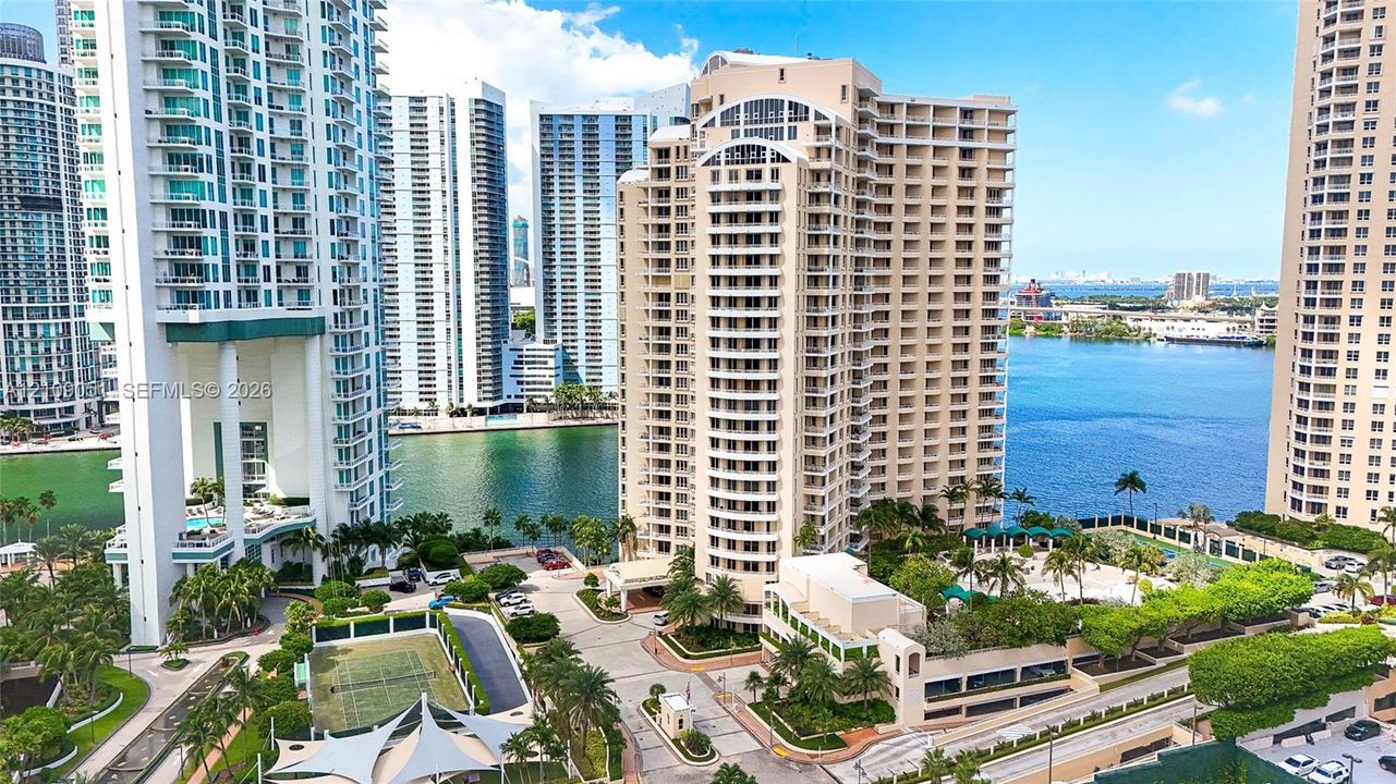 High-rise residential exterior featuring private balconies, tennis courts, a swimming pool, and gated entry, situated along the waterfront