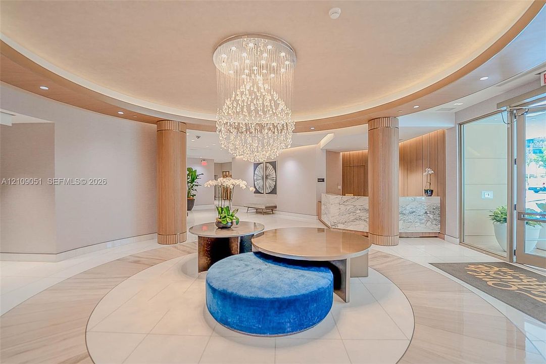 Grand foyer featuring a circular recessed ceiling, integrated crystal chandelier, polished stone flooring, custom wood-paneled pillars, and a stone-clad reception desk