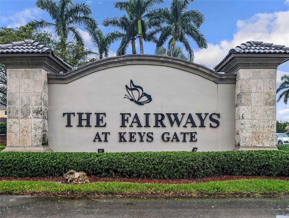 Monument-style neighborhood signage featuring stone-clad columns, arched stucco wall, and mature palm tree landscaping