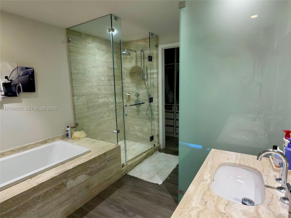 Bathroom featuring a soaking tub with stone-finish surround, glass-enclosed shower with adjustable rainfall showerhead, stone-finish vanity countertop, and frosted glass partitioning