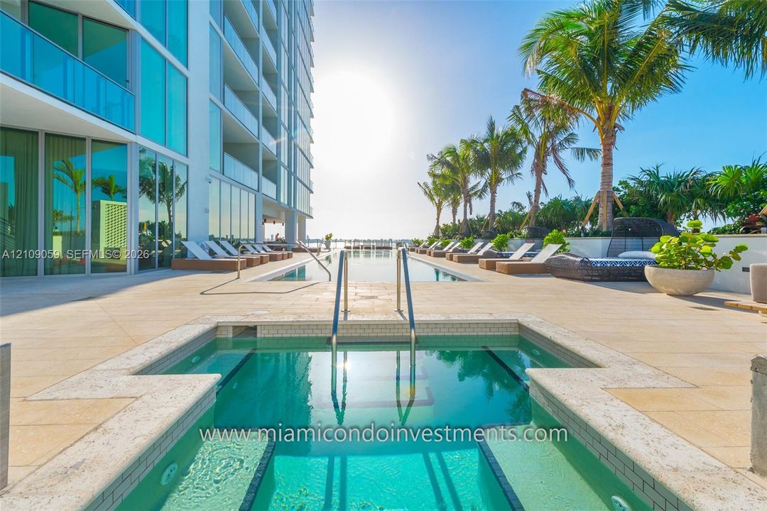 Pool deck featuring an in-ground spa, stone-finish paving, full-height glass building exterior, and palm tree landscaping