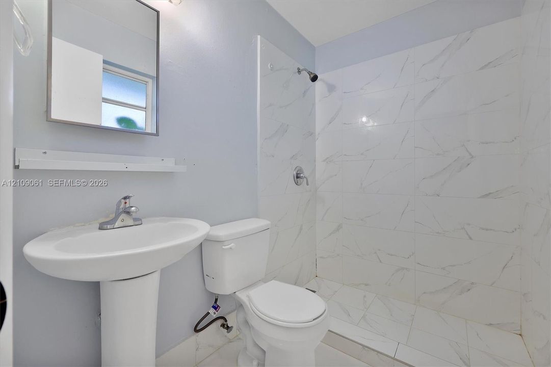 Bathroom featuring a walk-in shower with floor-to-ceiling stone-look tile, pedestal sink, chrome hardware, and rectangular wall-mounted mirror