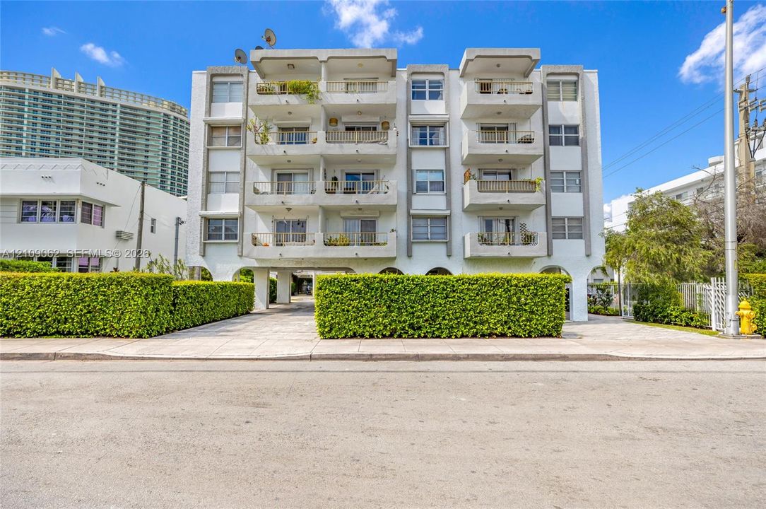Multi-story building exterior featuring a white stucco facade, stacked private balconies, arched ground-level entry, and manicured privacy hedges
