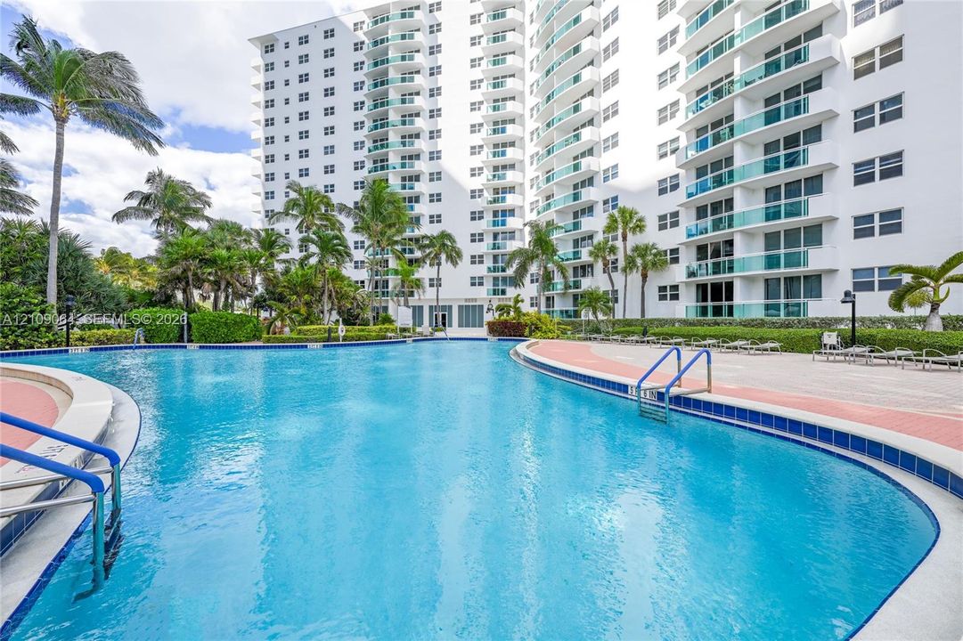 Swimming pool featuring a concrete deck with perimeter landscaping, metal handrails, and surrounding high-rise residential structure with private balconies