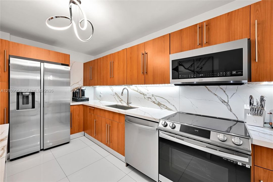 Kitchen featuring wood-finish cabinetry, stone-patterned backsplash, stainless steel appliances, under-cabinet lighting, and large-format tile flooring