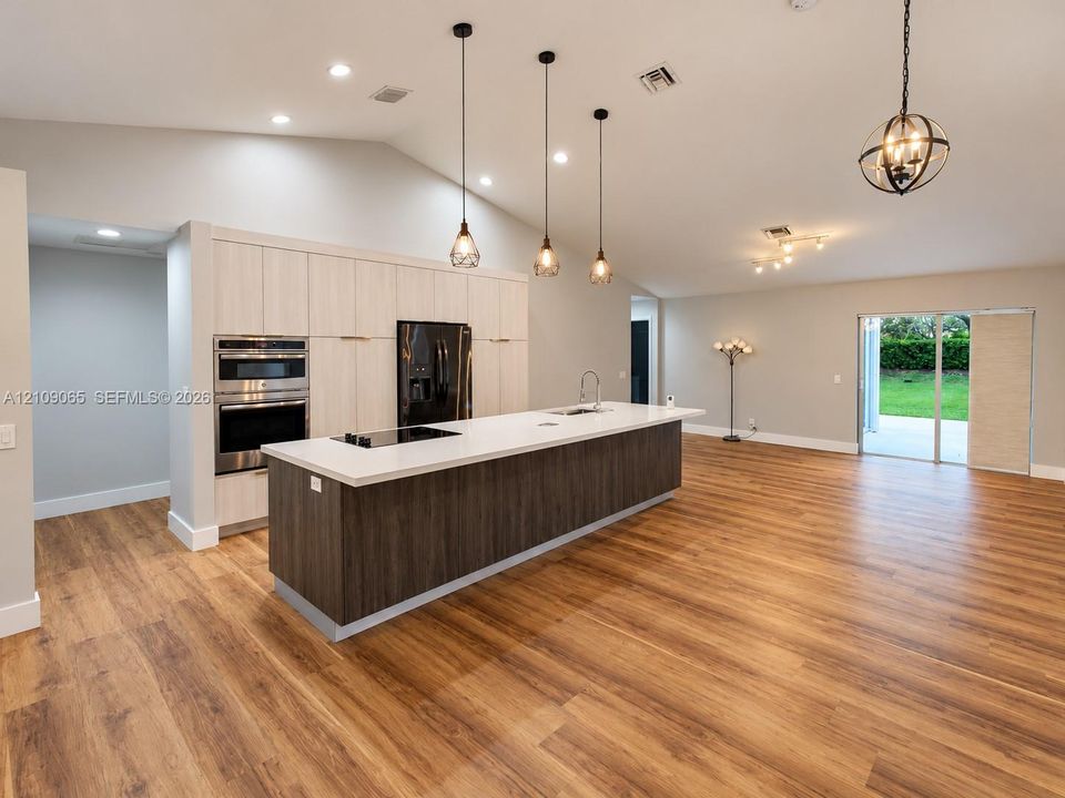 Open-concept kitchen featuring vaulted ceilings, wood-finish flooring, a central island with a solid surface countertop, light-toned cabinetry, and built-in double wall ovens