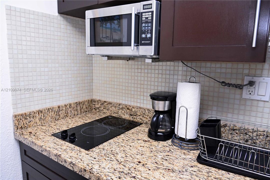 Kitchen area featuring dark cabinetry, stone-finish countertops, a two-burner electric cooktop, a stainless steel over-the-range microwave, and a mosaic tile backsplash