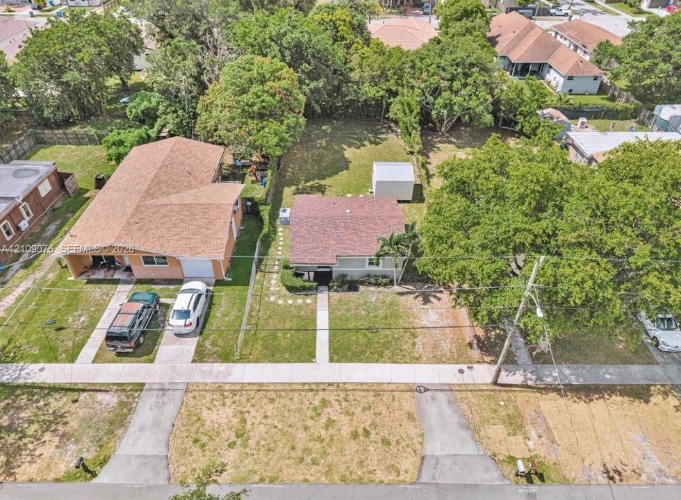Aerial perspective of residential lot featuring a paved concrete driveway, extensive grass lawn, mature trees, and a detached storage structure