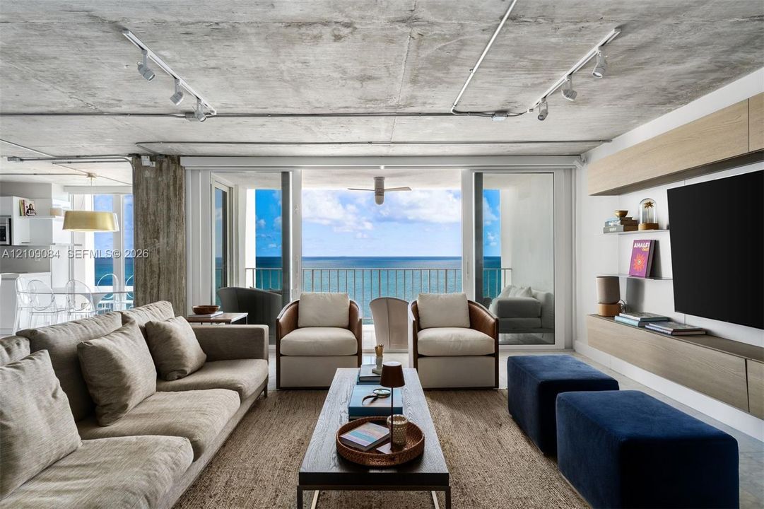 Exposed concrete ceiling with industrial track lighting, floor-to-ceiling glass doors, wall-mounted light wood-finish cabinetry, and balcony access featuring ocean water scenery