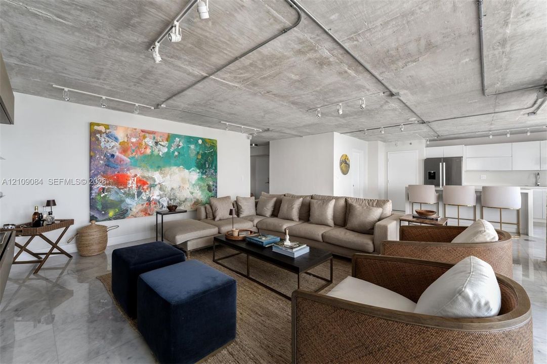 Contemporary kitchen featuring high-gloss white cabinetry, a waterfall-edge quartz island, stainless steel appliances, exposed concrete ceiling with industrial track lighting, and floor-to-ceiling windows with ocean scenery